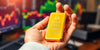 Hand holding a gold bar with a trading desk background.